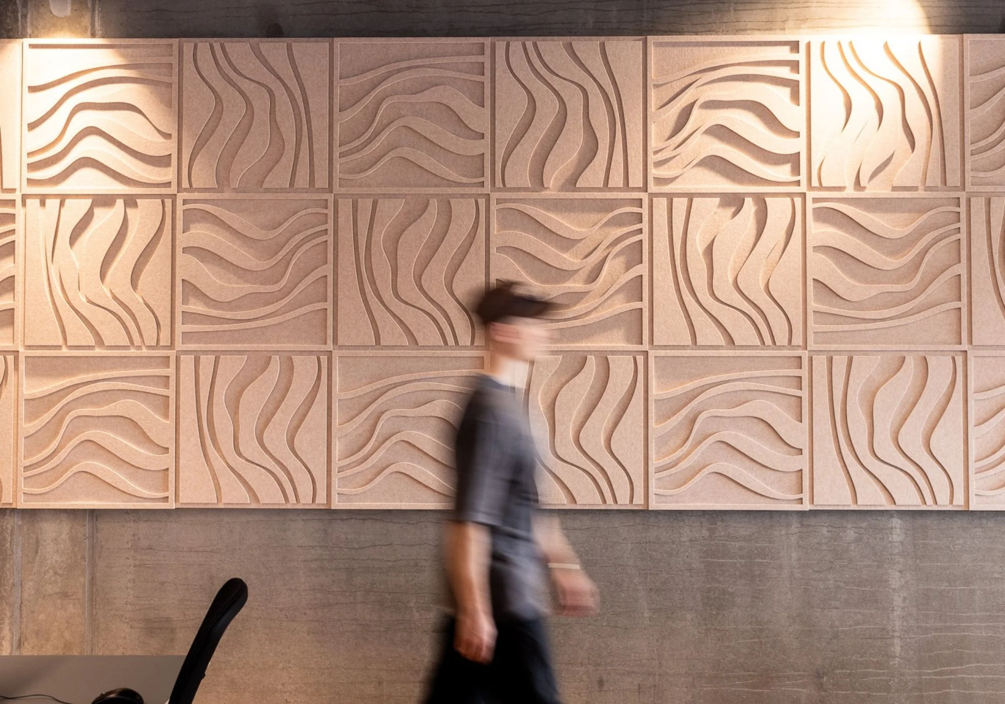 A blurred figure walking past a textured wave-patterned wall with soft lighting.