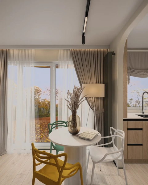 A cozy interior featuring a round white table surrounded by colorful chairs, with a large window allowing natural light to flood the space. There are decorative elements like a vase with dried flowers and soft curtains. The kitchen area can be seen in the background.