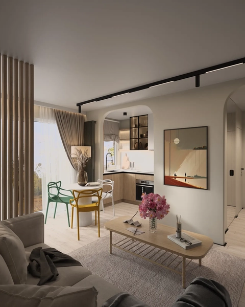 A modern living space featuring a dining area with a round table and colorful chairs, a small kitchen visible in the background, and large windows with natural light illuminating the room. Decorative touches include art on the walls and floral arrangements on the table.