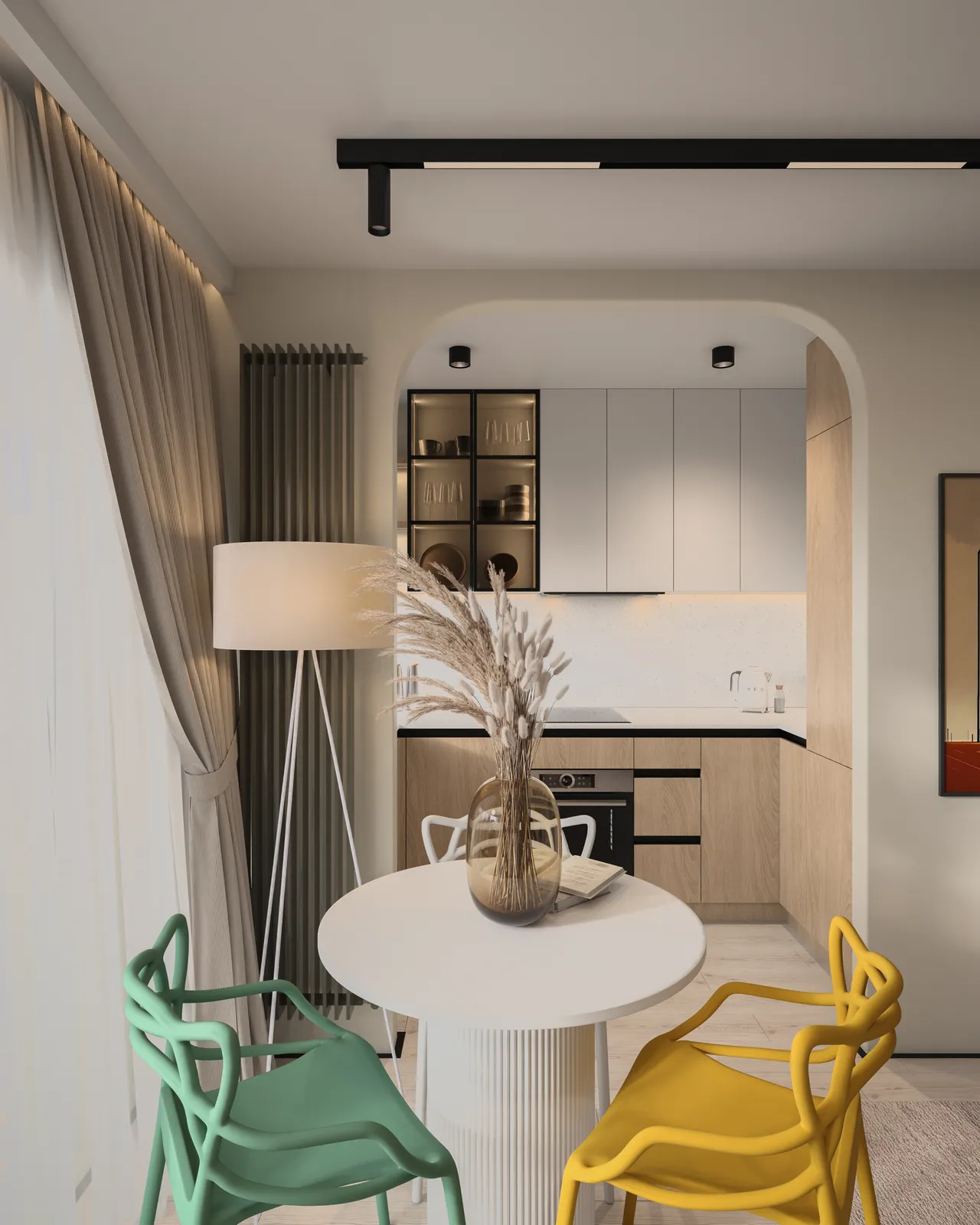 A modern kitchen with a small round dining table surrounded by colorful chairs, featuring a lamp and decorative plants. The kitchen has light cabinetry and dark countertops, with natural light filtering through sheer curtains.