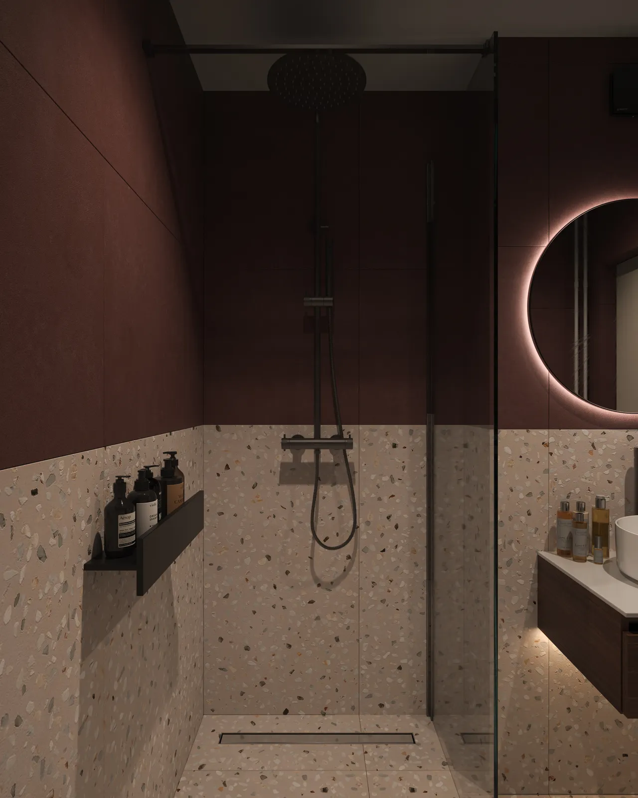 A modern bathroom featuring a walk-in shower with a sleek design, dark-colored walls, and terrazzo flooring. There are toiletries on a shelf, and ambient lighting is provided by a round mirror.