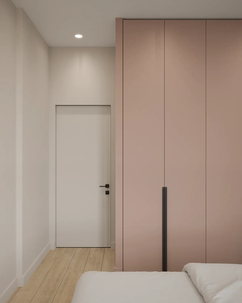 Minimalist bedroom interior featuring light wood floors, a white door, and a tall, blush pink built-in wardrobe with black handles, viewed from the foot of a white bed.