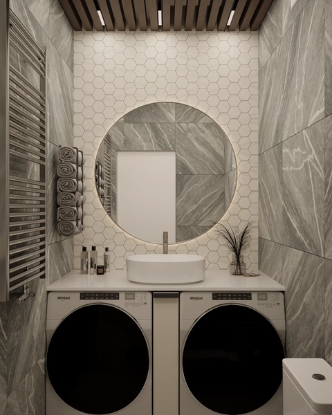 A modern bathroom featuring a circular mirror above a stylish sink, with a countertop. Below the sink, there are stacked laundry machines. The walls are covered with hexagonal tiles, and there is a towel rack on the side along with decorative items like plants and bottles. The ceiling has wooden slats with recessed lighting.