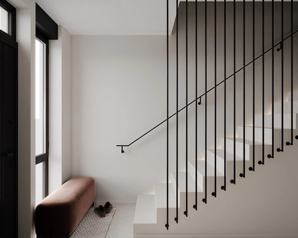 The entrance hall of the house, with a white staircase lit along the steps, a railing of vertical black rods, a bench upholstered in brick-colored velvet and a rug with a geometric pattern next to the black entrance door.