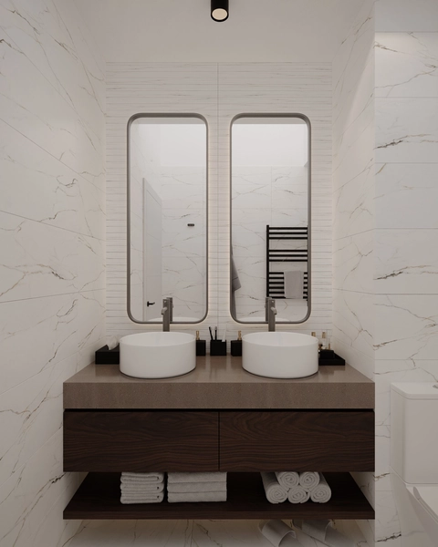 A bathroom clad in white marble, with two round washbasins on a stone countertop, wall-hung dark wood furniture, two illuminated vertical mirrors and rolled towels on the open shelf.