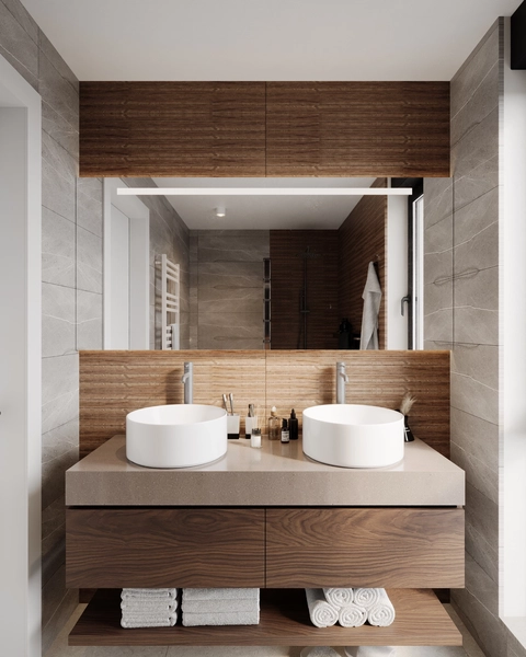 A bathroom with two round white washbasins on a stone countertop, wall-hung walnut furniture, a large illuminated mirror and a decorative fluted wood panel.