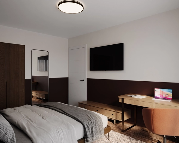 A bedroom with a double bed, a low wooden chest of drawers, a television mounted on the two-tone wall, a tall mirror and a desk with a caramel-colored upholstered chair.