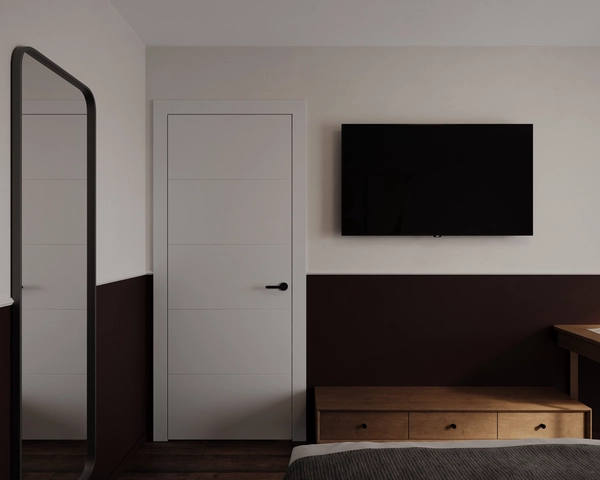 A detail of the bedroom, with a white door with a black handle, a tall framed mirror, a television on the two-tone wall and a wooden chest of drawers.