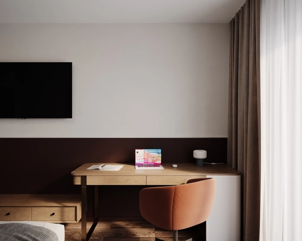 A wooden desk with a laptop and a table lamp, a brick-colored upholstered chair, a television mounted on the wall and beige drapes next to the window.