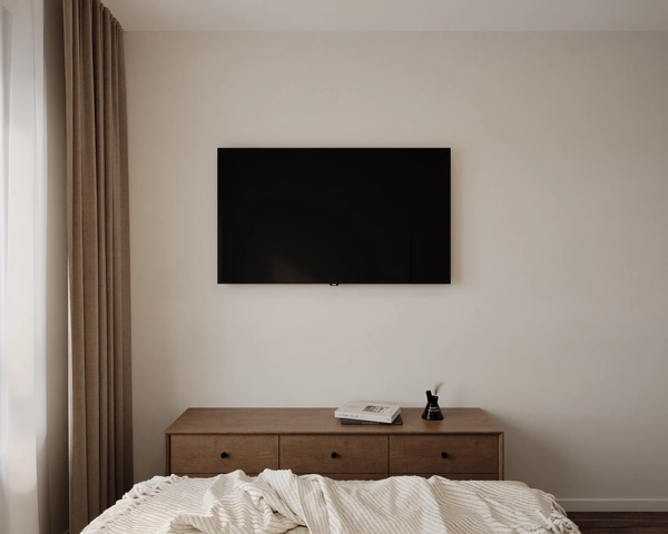 The TV wall of the bedroom, with a television mounted above a wooden chest of drawers and minimalist decor, against a warm beige background.