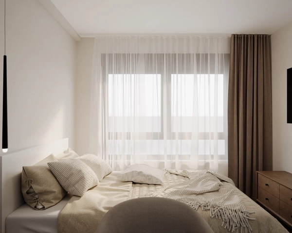 A bright bedroom, with a bed dressed in natural tones, a large window with sheer curtains and beige drapes and a wooden chest of drawers.