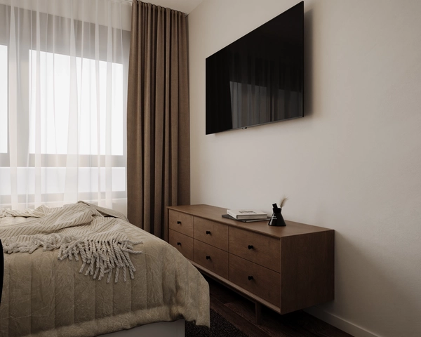 A bedroom with a television mounted on the wall, a long wooden chest of drawers, a bed with a textured throw and beige drapes next to the window.