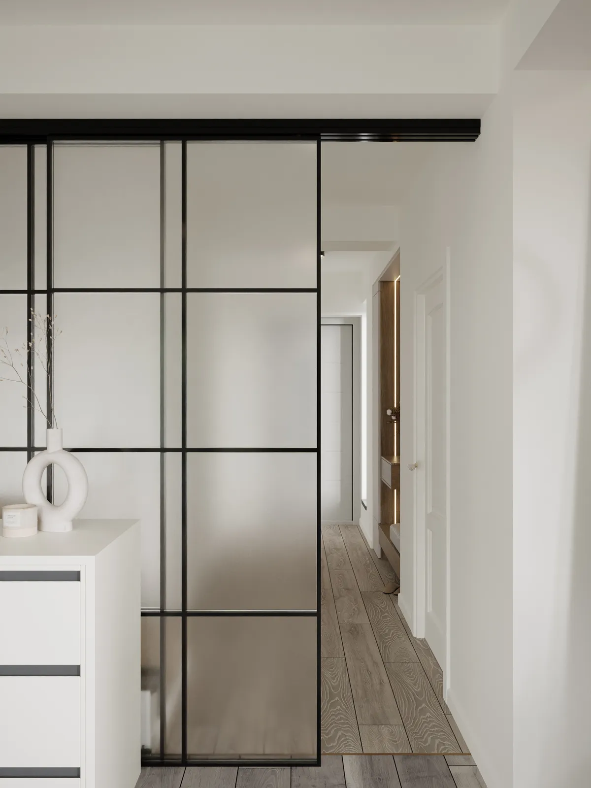 Minimalist hallway featuring a black-framed sliding frosted glass partition next to a white modern dresser and light wood flooring.